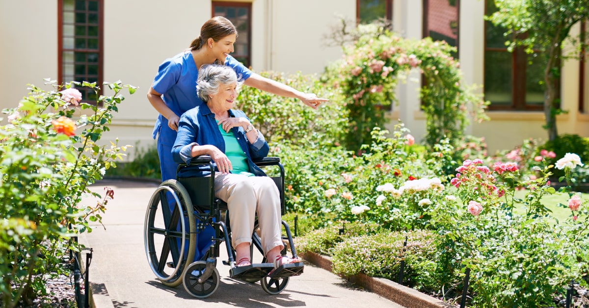 Cuidadora paseando con una mujer en silla de ruedas por un jardín, mientras señala las flores